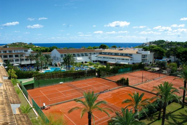 Ein Resort mit Tennispl&auml;tzen aus rotem Sand, Palmen, einem gro&szlig;en Poolbereich und wei&szlig;en Geb&auml;uden; im Hintergrund sind das Meer und ein klarer blauer Himmel zu sehen.