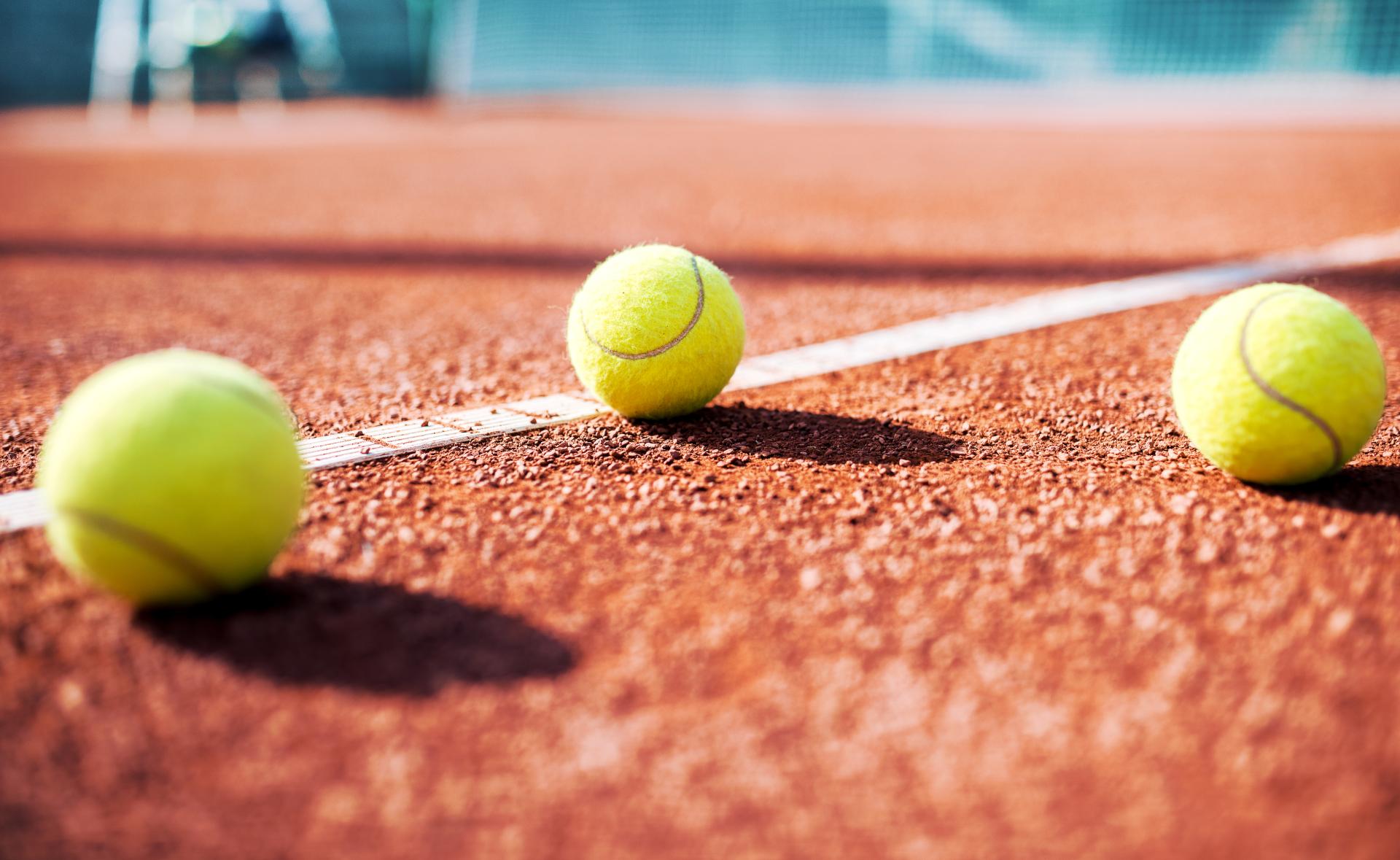 Drei gelbe Tennisbälle liegen auf der roten Sandplatz-Linie, scharf fokussiert im Vordergrund mit unscharfem Hintergrund.