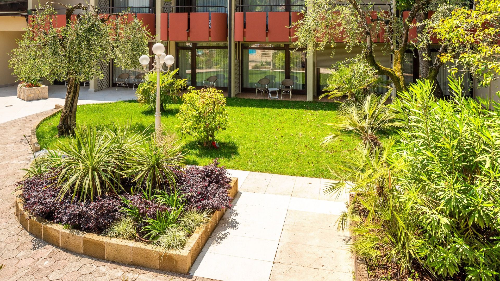 Ein Landschaftsgarten mit Gras, Bäumen und Sträuchern wird von einem Gehweg begrenzt. Terrassenstühle stehen vor den Glastüren des Sporthotels Olimpo mit roten Balkonplatten, die den Blick auf die nahe gelegenen Tennisplätze im Hintergrund freigeben.