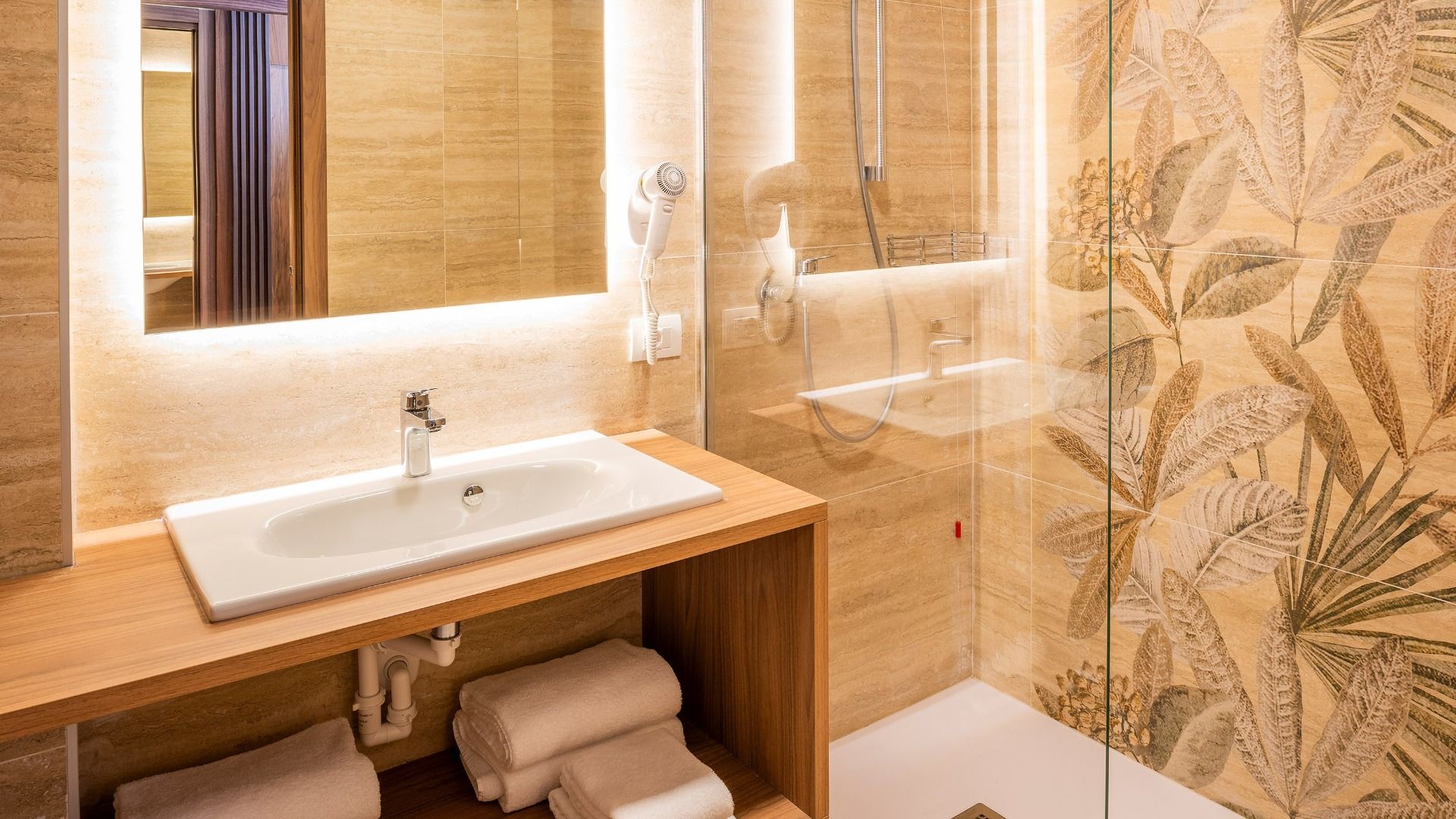 Das moderne Badezimmer im Sporthotel Olimpo in der Nähe des Gardasees verfügt über einen großen Spiegel, einen an der Wand befestigten Haartrockner, ein Waschbecken auf einem hölzernen Waschtisch mit Handtüchern darunter und eine verglaste Dusche mit gemu