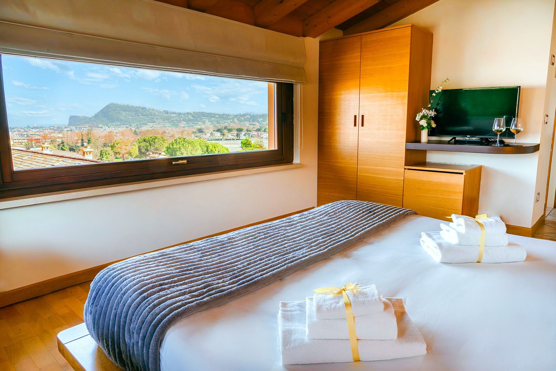 Ein gemütliches Zimmer im Hotel Donna Silvia verfügt über ein gemachtes Bett, zwei Handtuch-Sets, die mit gelben Bändern zusammengebunden sind, einen Kleiderschrank, einen Fernseher und ein großes Fenster mit atemberaubendem Blick auf die Stadt und die Be