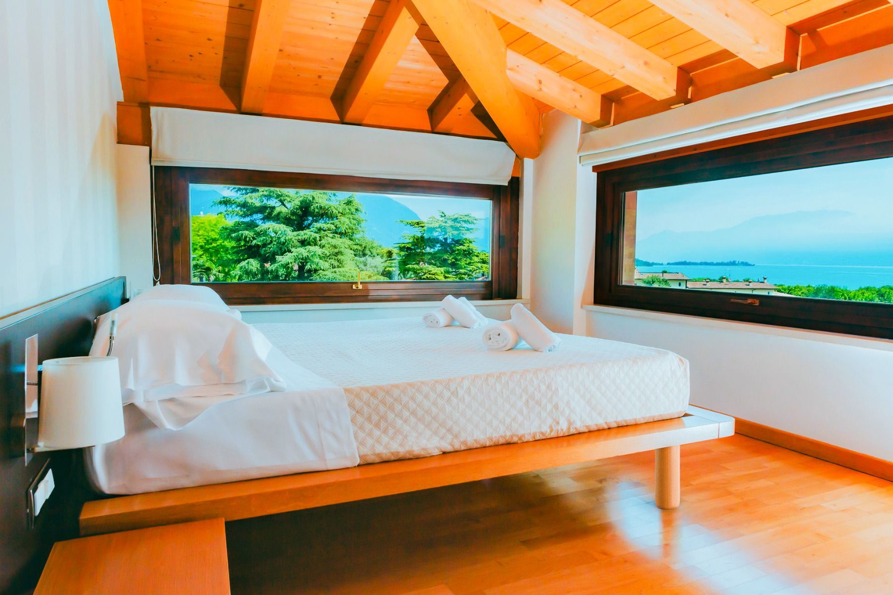 Ein helles Schlafzimmer mit Holzdecke, großen Fenstern, die einen malerischen Blick auf Bäume und Wasser bieten, einem ordentlich gemachten Bett mit weißer Bettwäsche und darauf aufgerollten Handtüchern – ein einladender Rückzugsort im Hotel Donna Silvia,