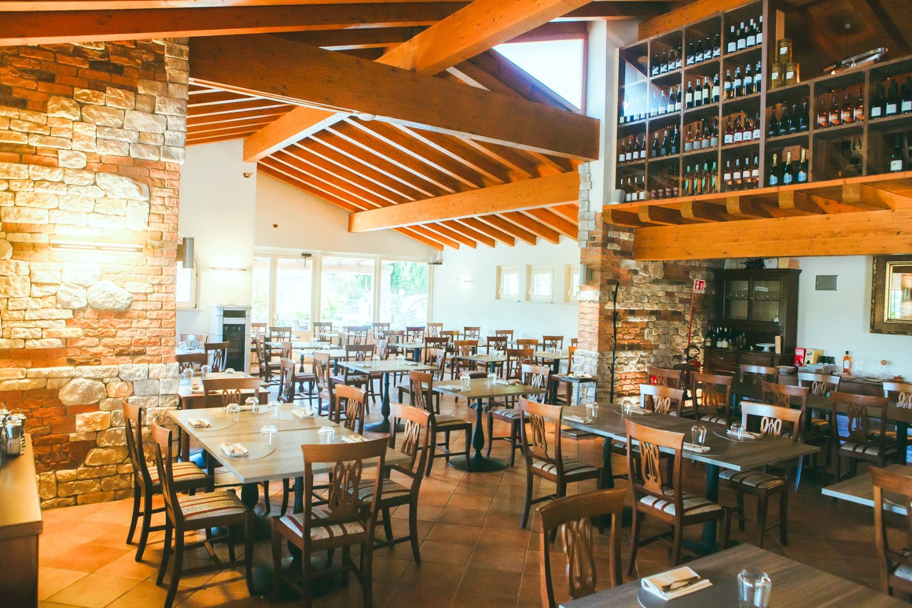 Ein helles, geräumiges Restaurant mit Holzbalken, Steinwänden sowie Tischen und Stühlen aus Holz. Auf hohen Regalen stehen Weinflaschen, und durch die großen Fenster strömt Sonnenlicht herein – ein perfekter Ort im Hotel Donna Silvia für Gäste, die nach e