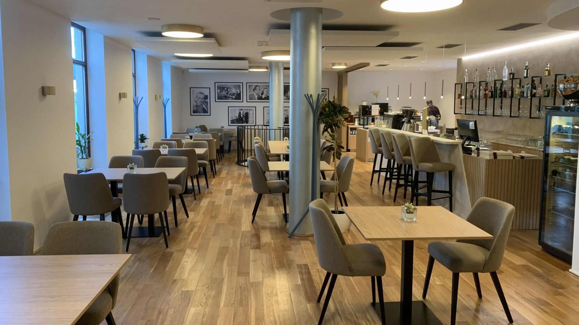 Das Hotel Biograf besticht durch eine moderne Café-Einrichtung mit hellen Holzböden, beigen Polstersesseln und quadratischen Tischen. Große Fenster lassen viel Licht herein, während an der Bar auf der rechten Seite Getränke und Desserts angeboten werden.
