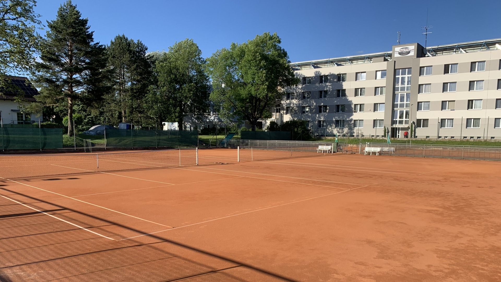Das Hotel Biograf verfügt über einen Sandtennisplatz mit Netzen und Bänken, der von Bäumen und einem grünen Zaun umgeben ist und unter einem strahlend blauen Himmel liegt. Im Hintergrund erhebt sich ein großes weißes Gebäude mit zahlreichen Fenstern.