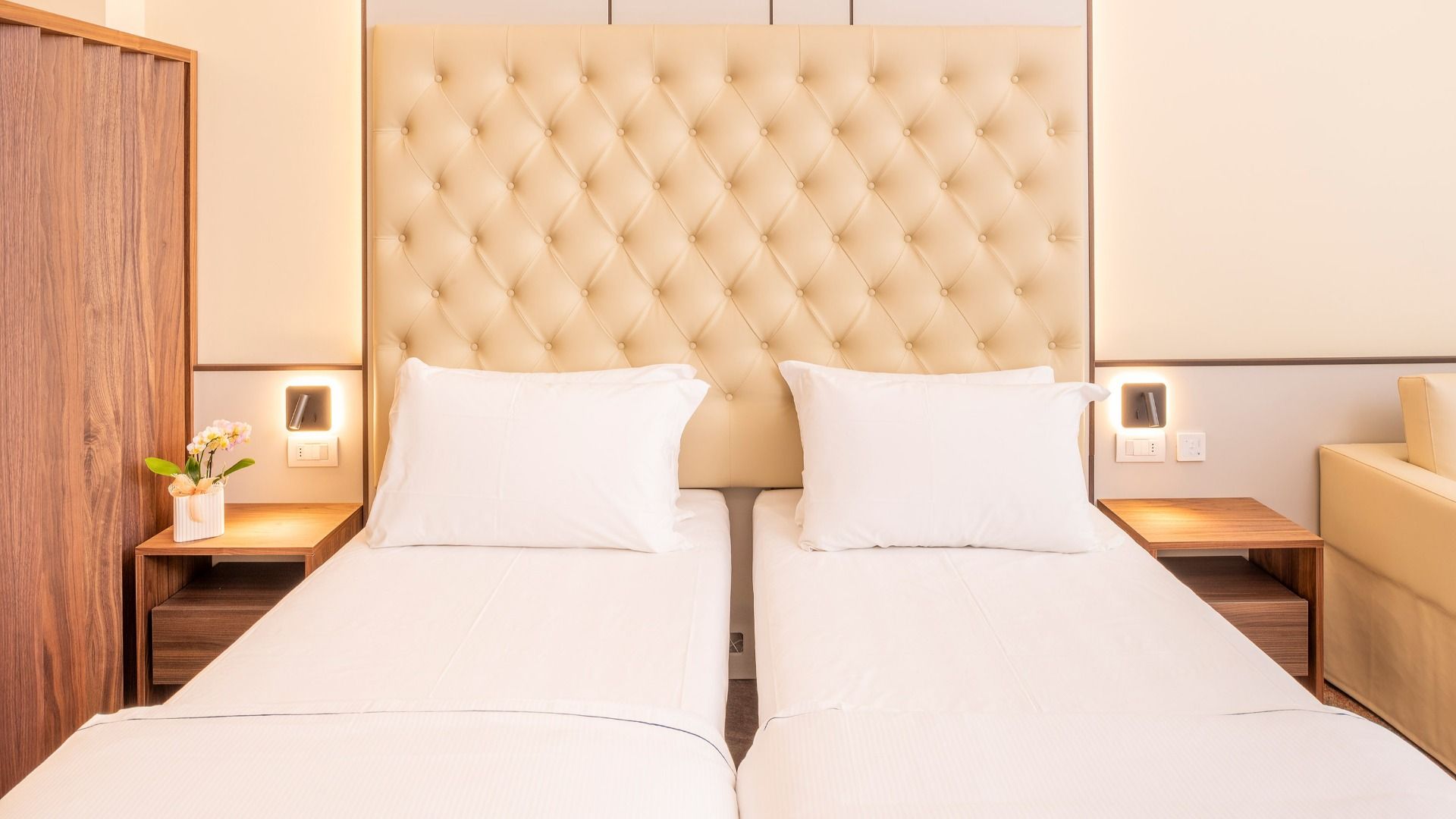 Zwei Einzelbetten mit weißer Bettwäsche und einem getufteten beigen Kopfteil, flankiert von hölzernen Nachttischen und Blumen, in einem hellen, modernen Zimmer im Sporthotel Olimpo in der Nähe des Gardasees, mit einfachem Zugang zu den Tennisplätzen.