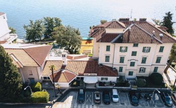 Luftaufnahme des Hotels Lovran mit roten Ziegeldächern, geparkten Autos davor, Bäumen und einem See im Hintergrund an einem sonnigen Tag.