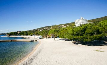 Hinter üppig grünen Bäumen und Sträuchern erhebt sich das hohe, moderne, weiße Gebäude des Hotels Omorika. Eine lange Steintreppe führt hinauf zum Eingang, an dessen Vorderseite ein blaues „Hotel Omorika“-Schild angebracht ist.