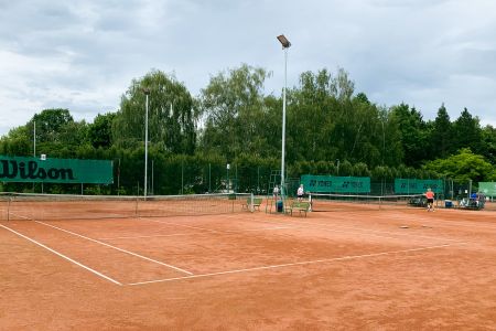 Der TJ Jiskra Třeboň-Sandtennisplatz befindet sich im Freien und ist von grünen Bäumen und einem Zaun umgeben. In der Nähe stehen Menschen, deren Tennisausrüstung zu sehen ist. Unter einem bewölkten Himmel wirkt die Szene ruhig und friedlich.