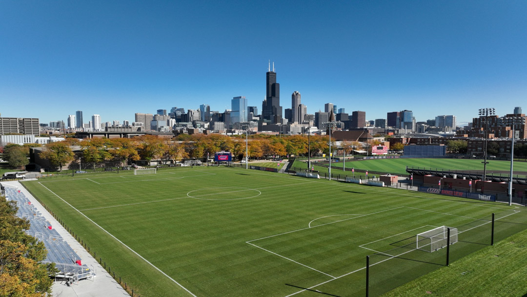 Fu&szlig;ballfeld mit gepflegtem Rasen vor einer modernen Skyline, umgeben von Flutlichtmasten und Trib&uuml;nen unter klarem, blauem Himmel.