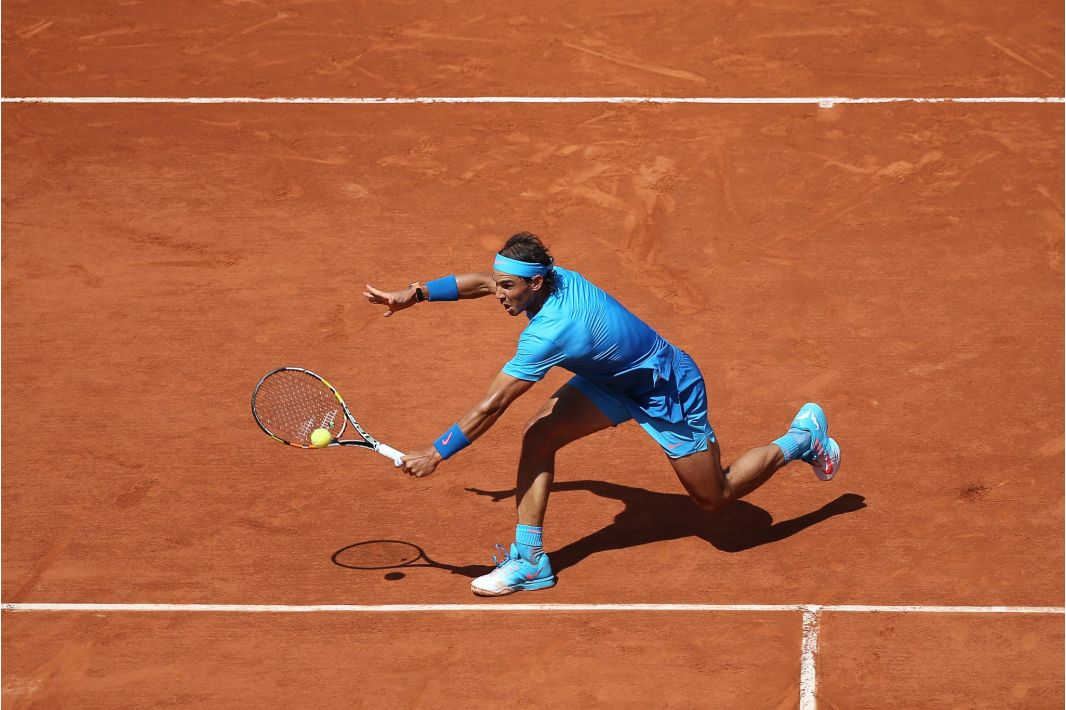 Ein Tennisspieler in einem blauen Outfit streckt sich, um auf einem roten Sandplatz einen Vorhandschlag zu machen, wobei er einen Fuß vom Boden abhebt und den Blick auf den Ball richtet.