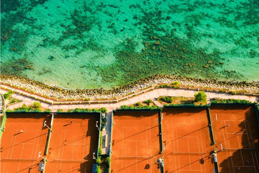 Luftaufnahme von Tennisplätzen aus rotem Sand, die von einem Steinweg begrenzt werden, mit türkisfarbenem, flachem Meerwasser und felsigem Ufer am oberen Bildrand.