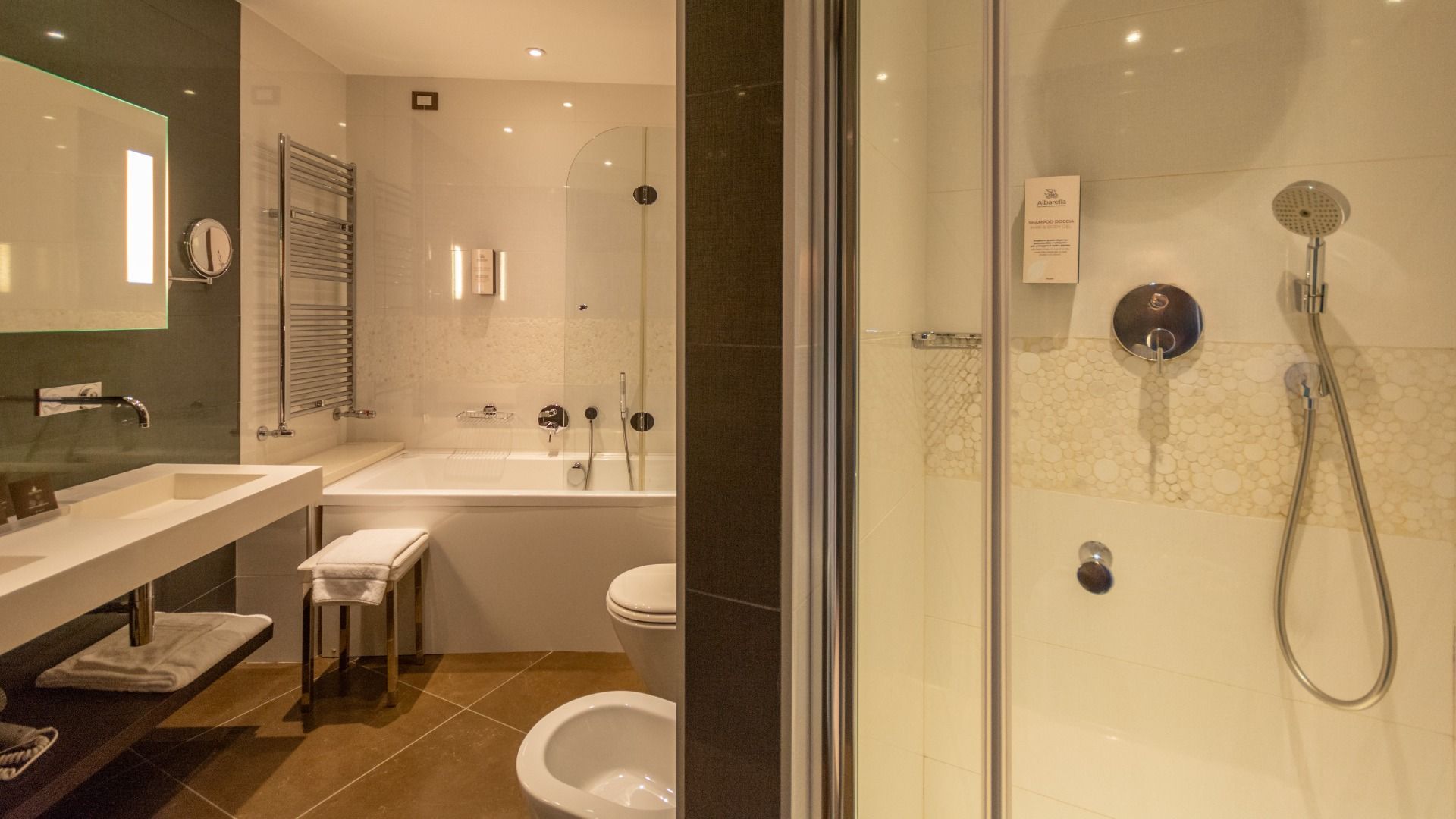 Modernes Badezimmer im Albarella Golf Hotel mit Glasduschkabine, wandmontiertem Duschkopf, Badewanne, Waschbecken und Spiegel, Handtuchhalter, neutralen Fliesen und Beleuchtung – ideal für entspannende Erholung nach dem Sport in ruhiger Atmosphäre.