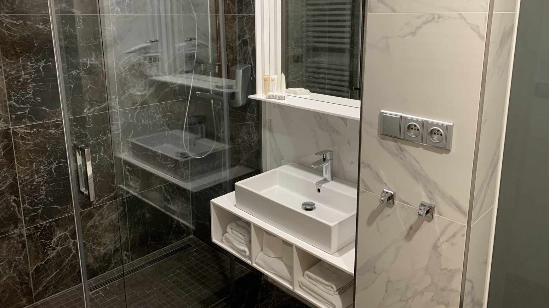 Das Badezimmer im Hotel Biograf verfügt links über eine Glasdusche, in der Mitte über ein weißes Waschbecken mit einem Spiegelschrank darüber sowie darunter über Ablagen, auf denen gefaltete Handtücher liegen. Dunkle und helle Marmorfliesen zieren die Wän