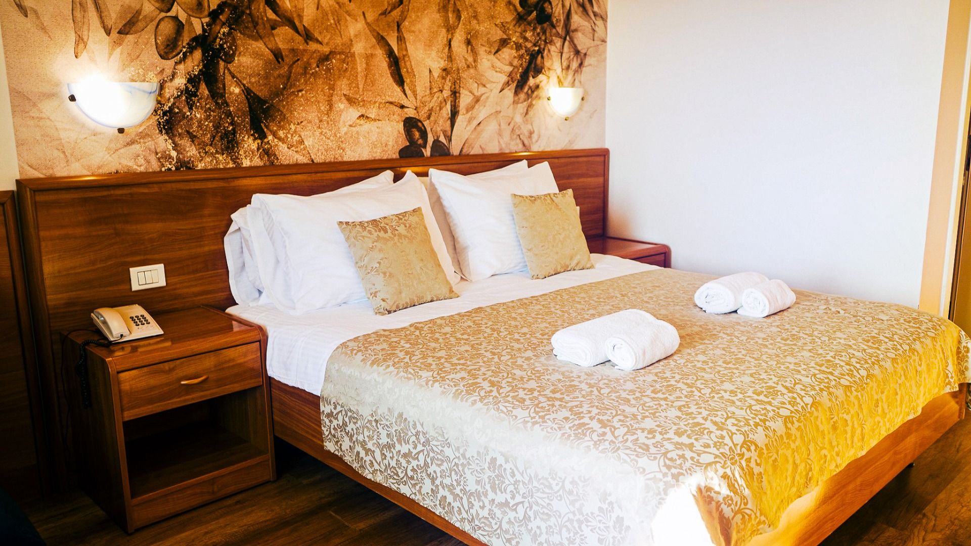 Ein ordentlich gemachtes Bett im Hotel Villa Letan ist mit einem golden gemusterten Bezug, zwei weißen Handtuchrollen, dekorativen Kissen und einem hölzernen Kopfteil mit sanften Wandlampen ausgestattet - ein gemütliches Ambiente und ein ausgezeichnetes P