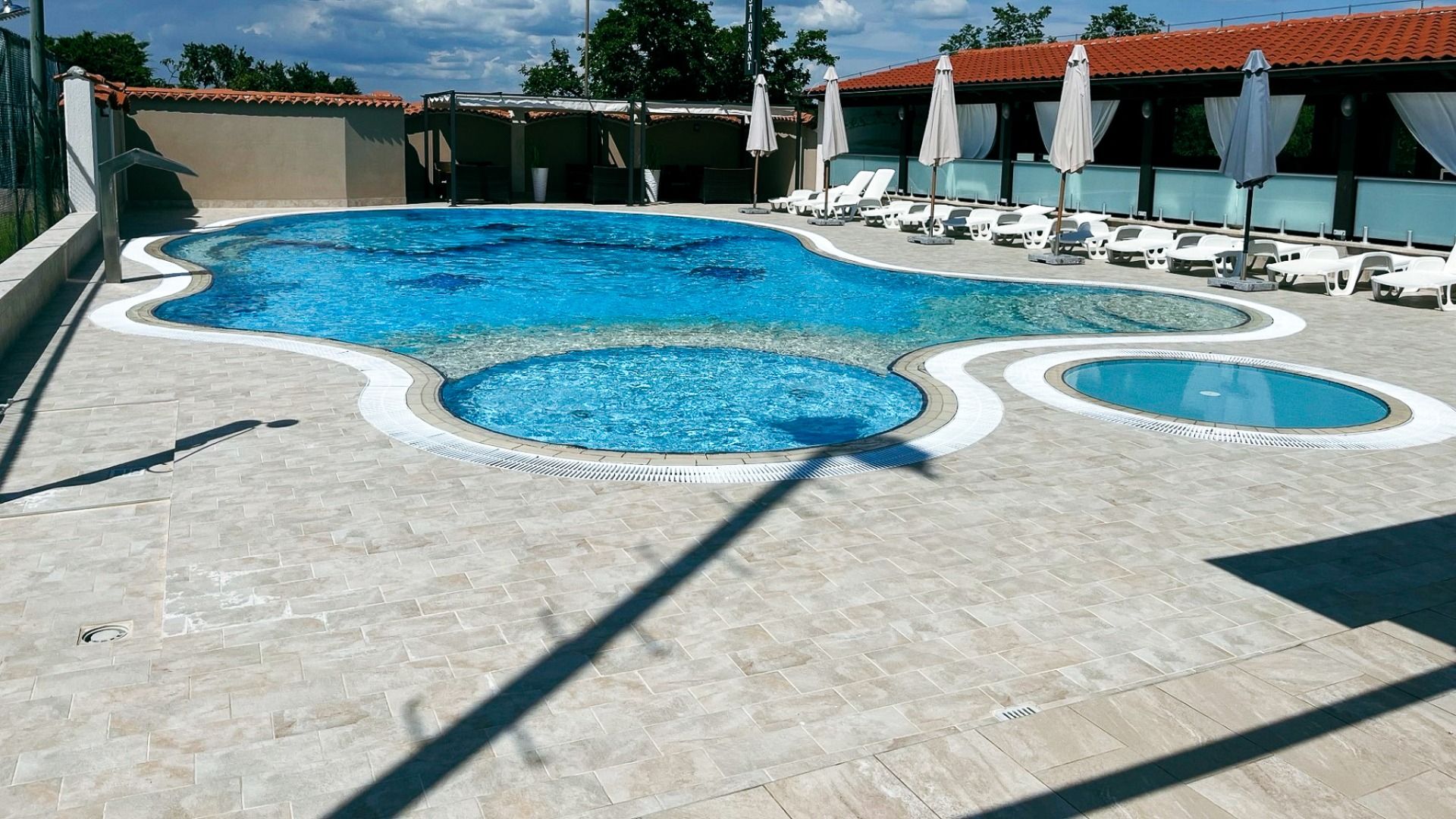 Im Hotel Villa Letan bildet ein moderner, geschwungener Außenpool das Herzstück des Wellnessbereichs, der von einer gefliesten Terrasse mit weißen Liegestühlen und Sonnenschirmen begrenzt wird. Bäume und Gebäude bieten eine malerische Kulisse unter einem 