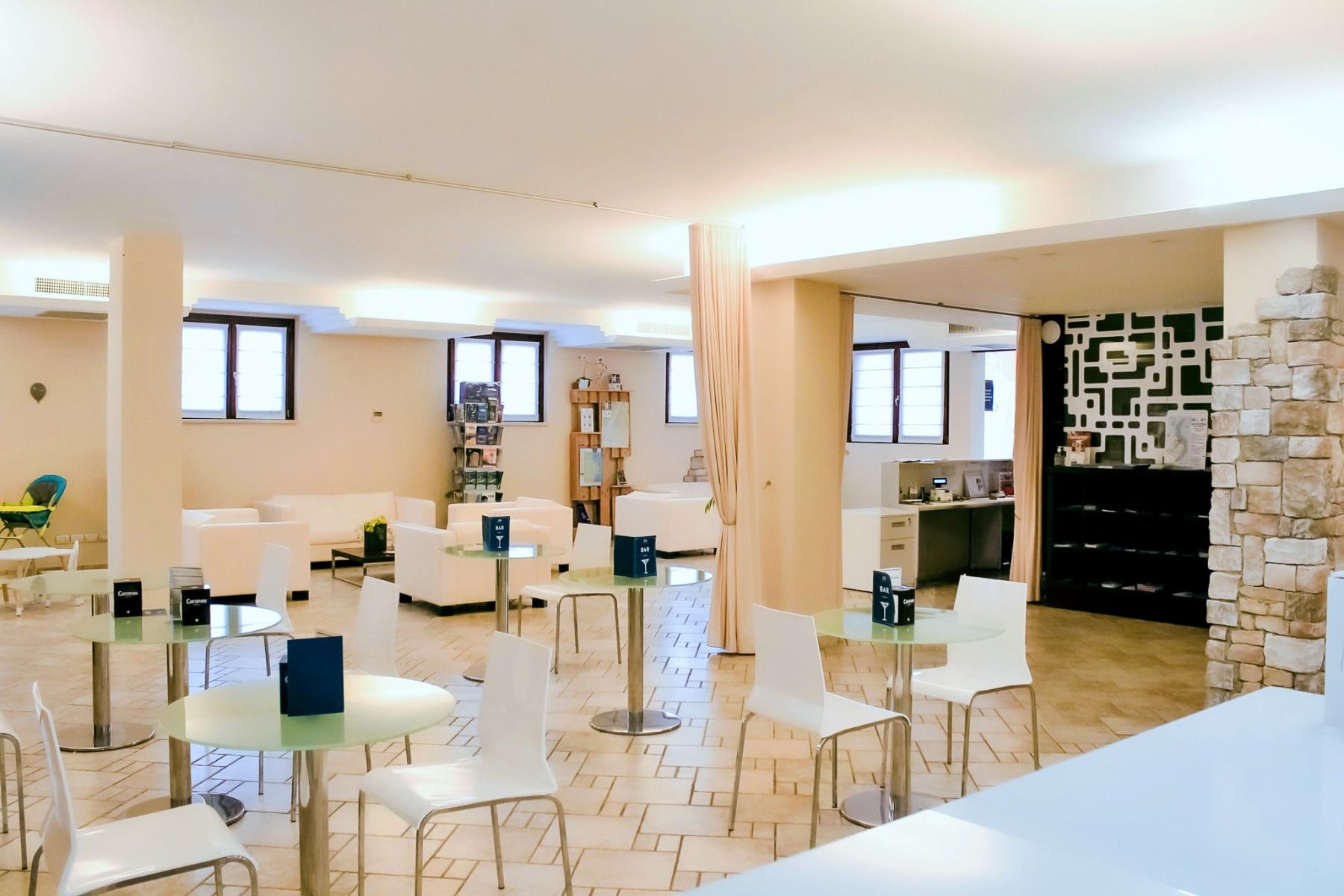 Die moderne Lounge im 4-Sterne-Hotel Donna Silvia besticht durch weiße Stühle und Tische, eine Akzentwand aus Stein und sanfte Beleuchtung. Regale und Dekoration sind gut sichtbar, und die Fenster lassen Tageslicht herein, wodurch eine klare und einladend