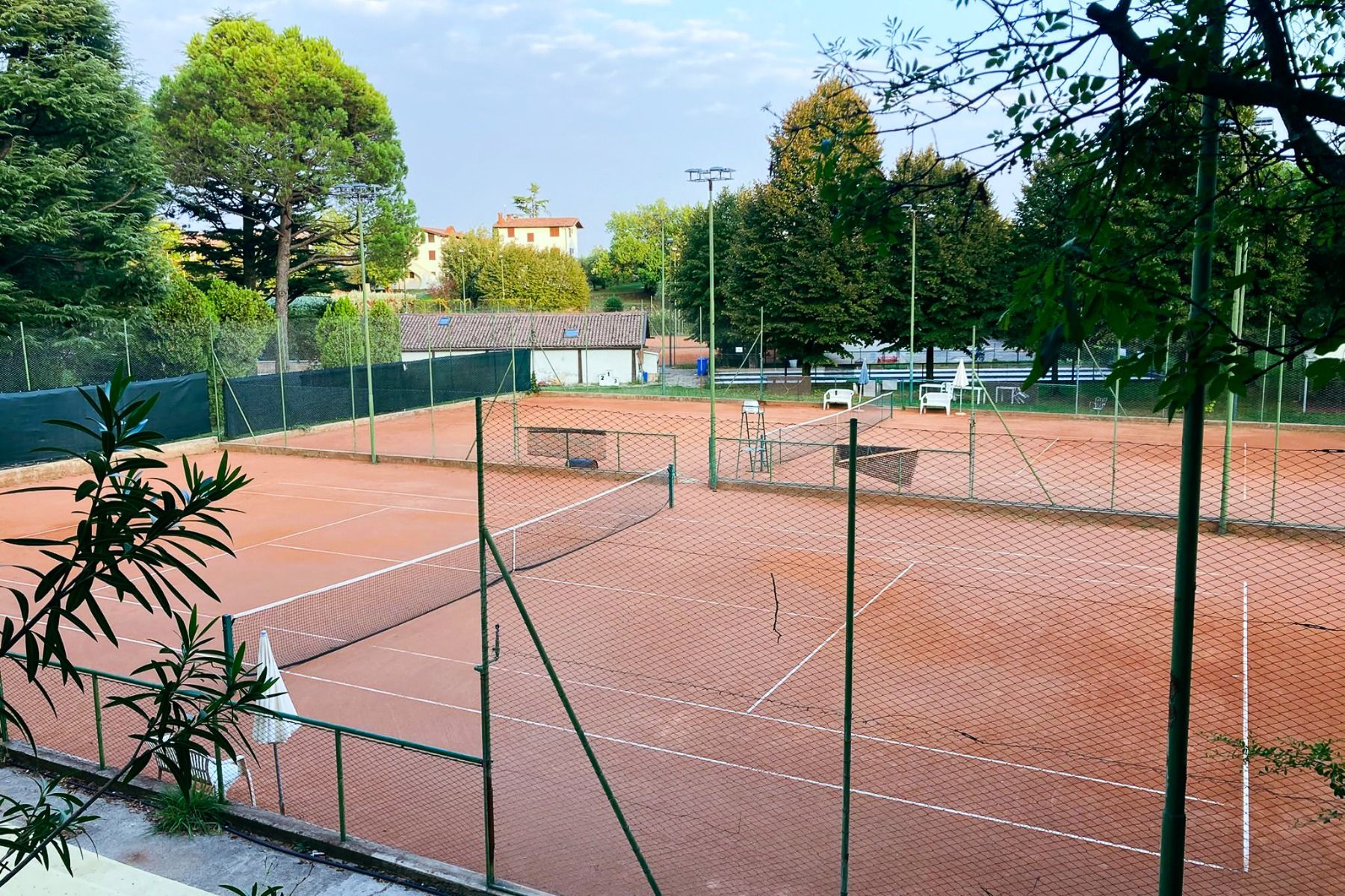 Ein Sandtennisplatz, umgeben von einem grünen Zaun, vor dem sich Bäume und Gebäude abzeichnen. Der leere Platz verfügt auf der rechten Seite über Sitzgelegenheiten im Freien und bietet damit den idealen Rahmen für ein Tennistrainingslager im 4-Sterne-Hote