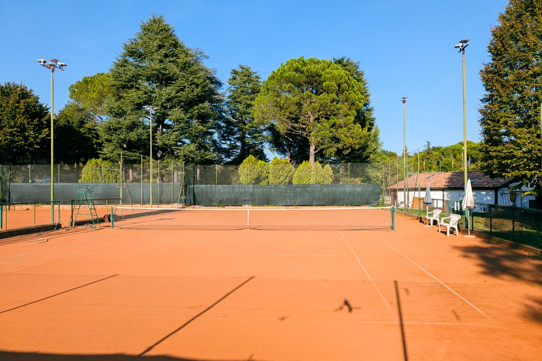Sandtennisplatz am Hotel Donna Silvia mit Netz, hohen Lichtmasten und umliegenden Bäumen.