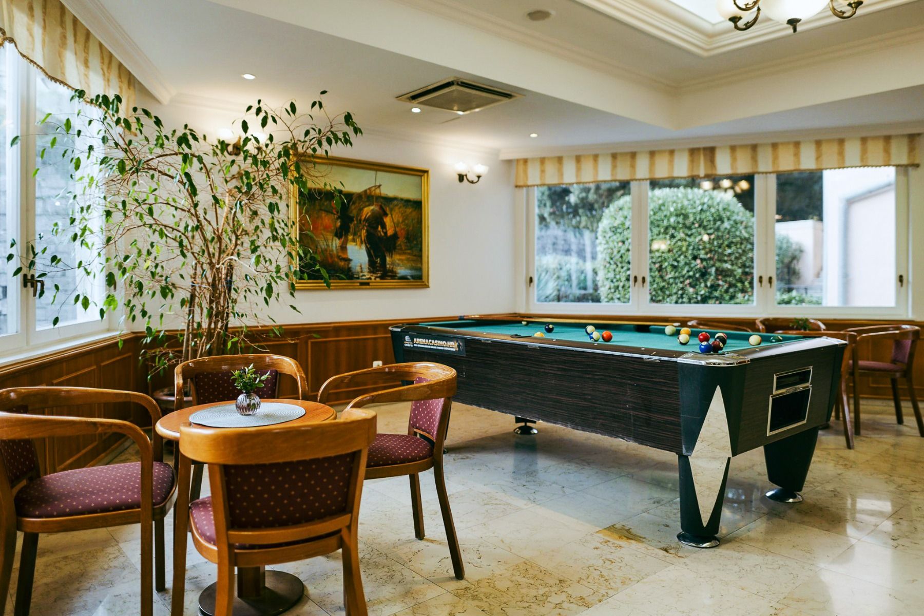 Genießen Sie im Hotel Lovran ein gemütliches Spielzimmer mit einem Billardtisch, vier Stühlen mit roten Polstern, die um einen kleinen runden Tisch herum angeordnet sind, großen Fenstern, durch die Tageslicht hereinströmt, einer Topfpflanze für etwas Grün
