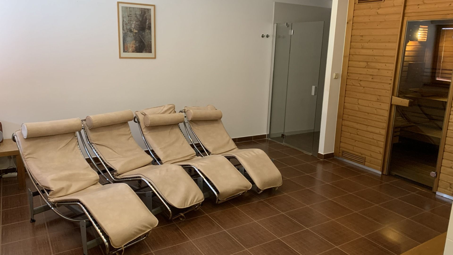 Vier beigefarbene Liegestühle stehen nebeneinander in dem gefliesten Raum des Hotels Biograf; rechts davon befindet sich eine Holzsauna, hinten eine Duschkabine mit Glastür und an der weißen Wand hängt ein gerahmtes Bild.