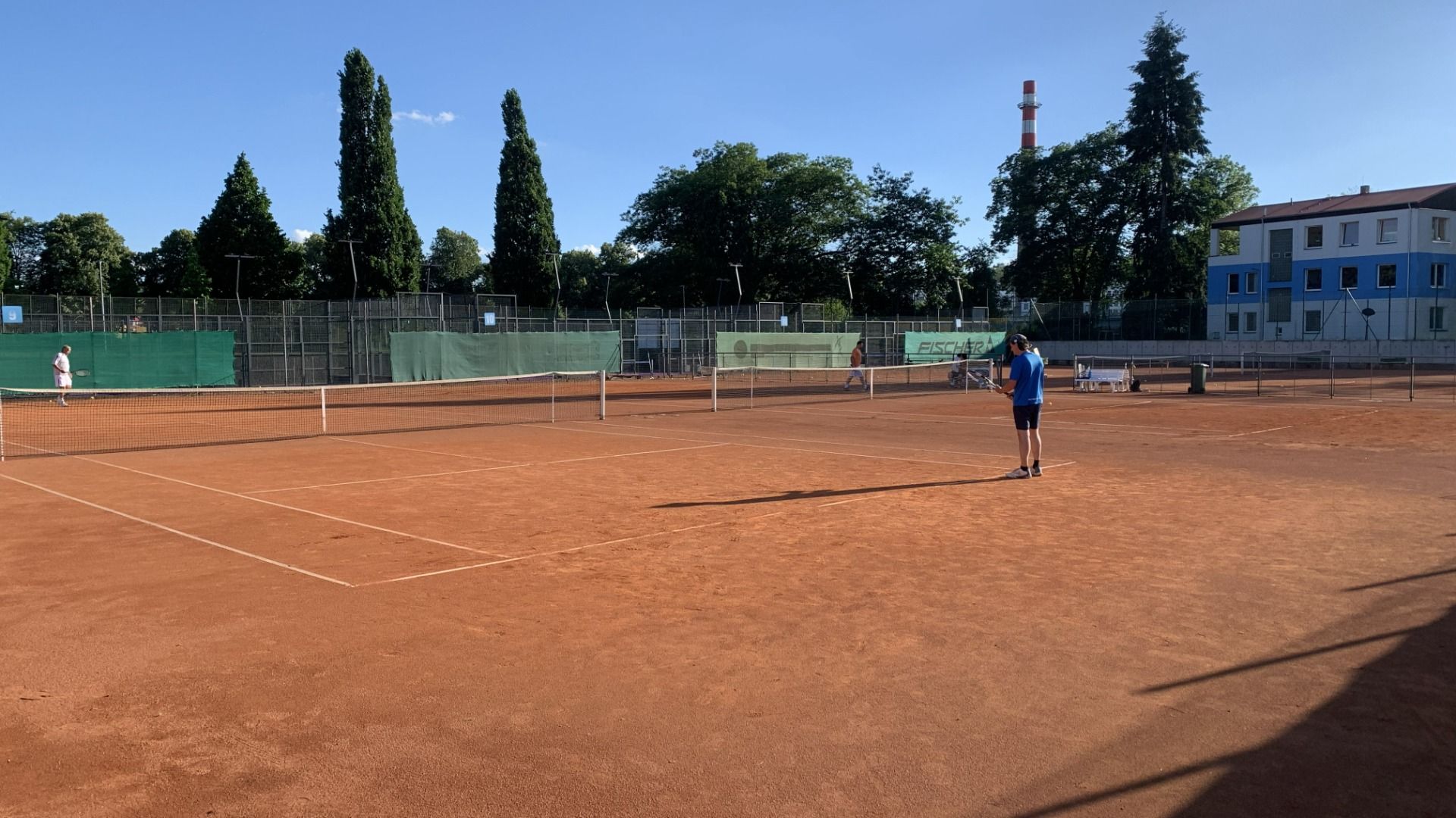 Drei Personen spielen unter strahlend blauem Himmel auf einem Sandplatz beim Hotel Biograf Tennis. Hohe Bäume, ein umzäuntes Gelände und ein Gebäude bilden den Hintergrund, während sich die Schatten der Spieler über den Platz ziehen.