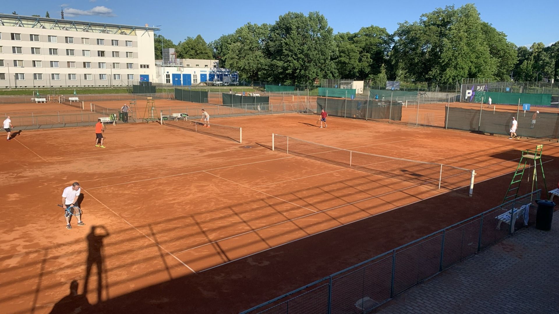 Im Hotel Biograf können die Gäste unter freiem Himmel auf mehreren orangefarbenen Sandplätzen Tennis spielen. Bäume, ein Gebäude und eine Umzäunung umgeben die Plätze, während lange Nachmittagsschatten eine entspannte Atmosphäre schaffen.