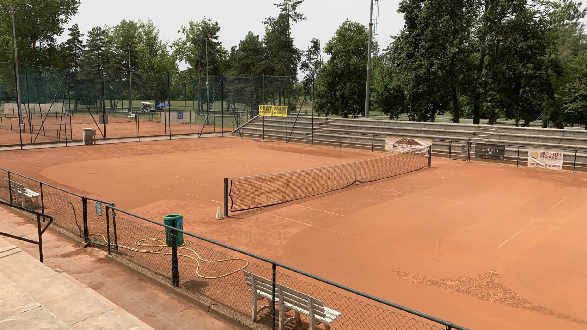 Das Albarella Golf Hotel verfügt über einen leeren Sandtennisplatz mit einem Netz in der Mitte, grünen Zäunen, Bänken und Bäumen – und bietet so eine ruhige Atmosphäre zur Entspannung nach dem Sport. Im bewölkten Hintergrund sind Tribünen und Banner zu se