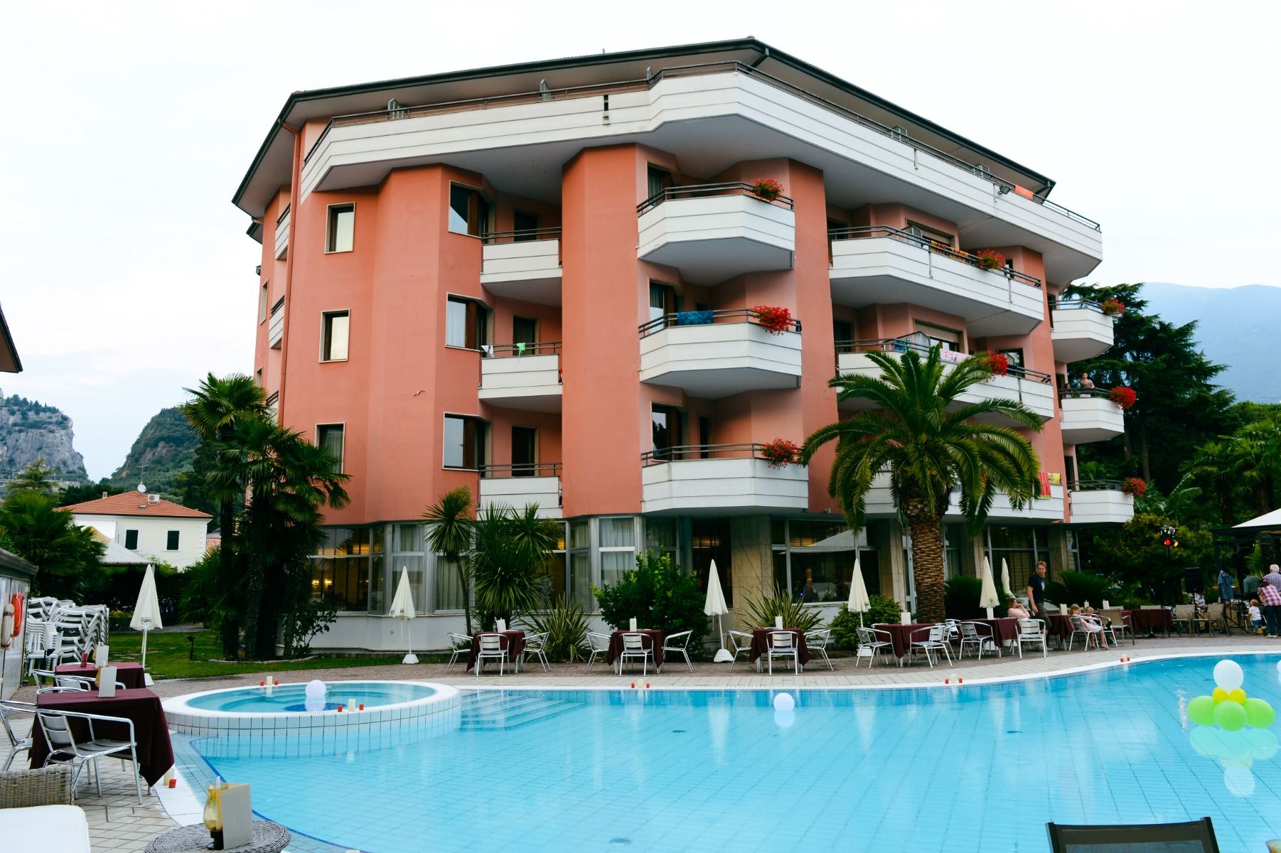 Hotelgebäude mit Pool, Sonnenliegen, Palmen und Balkonen, im Hintergrund Berge.