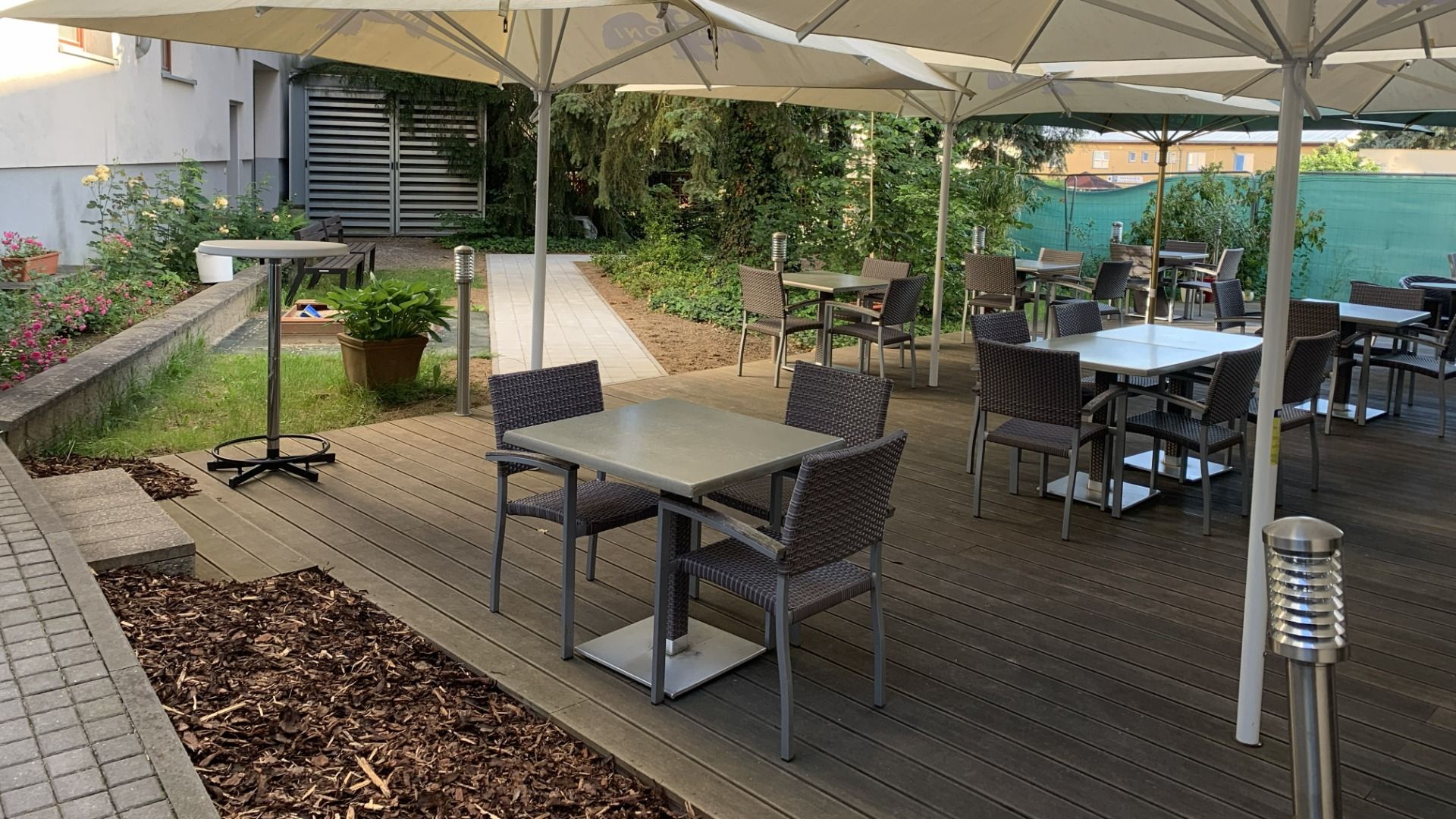 Die Außenterrasse des Hotels Biograf verfügt über mehrere Tische und Stühle unter großen weißen Sonnenschirmen, die auf einer makellosen Holzterrasse stehen, umgeben von Grünpflanzen und Blumen, was eine ruhige und einladende Atmosphäre schafft.