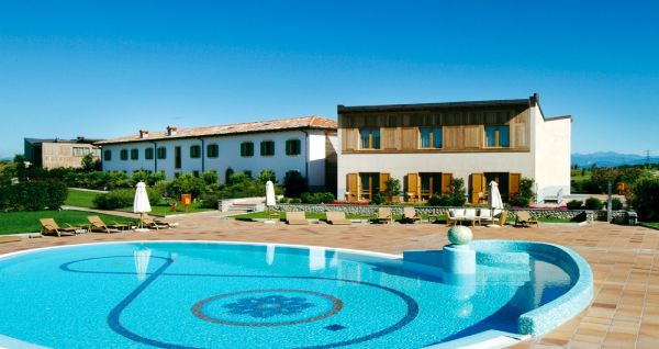 Rundpool mit blauer Mosaikverzierung, Sonnenliegen und Hotelgebäude mit weißer Fassade im Hintergrund.