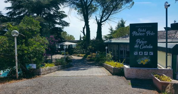 Eingangsbereich des Hotels Marco Polo, von Bäumen und Pflanzen umgeben, mit einem Weg zur Rezeption und einem großen Schild mit Hotelnamen.