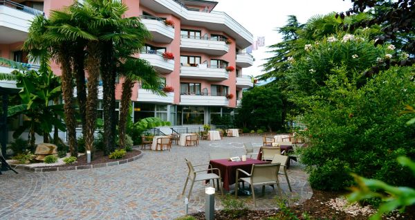 Außenbereich eines Hotels mit rosa Fassade, Balkonen und Palmen, sowie Sitzgelegenheiten im Garten.