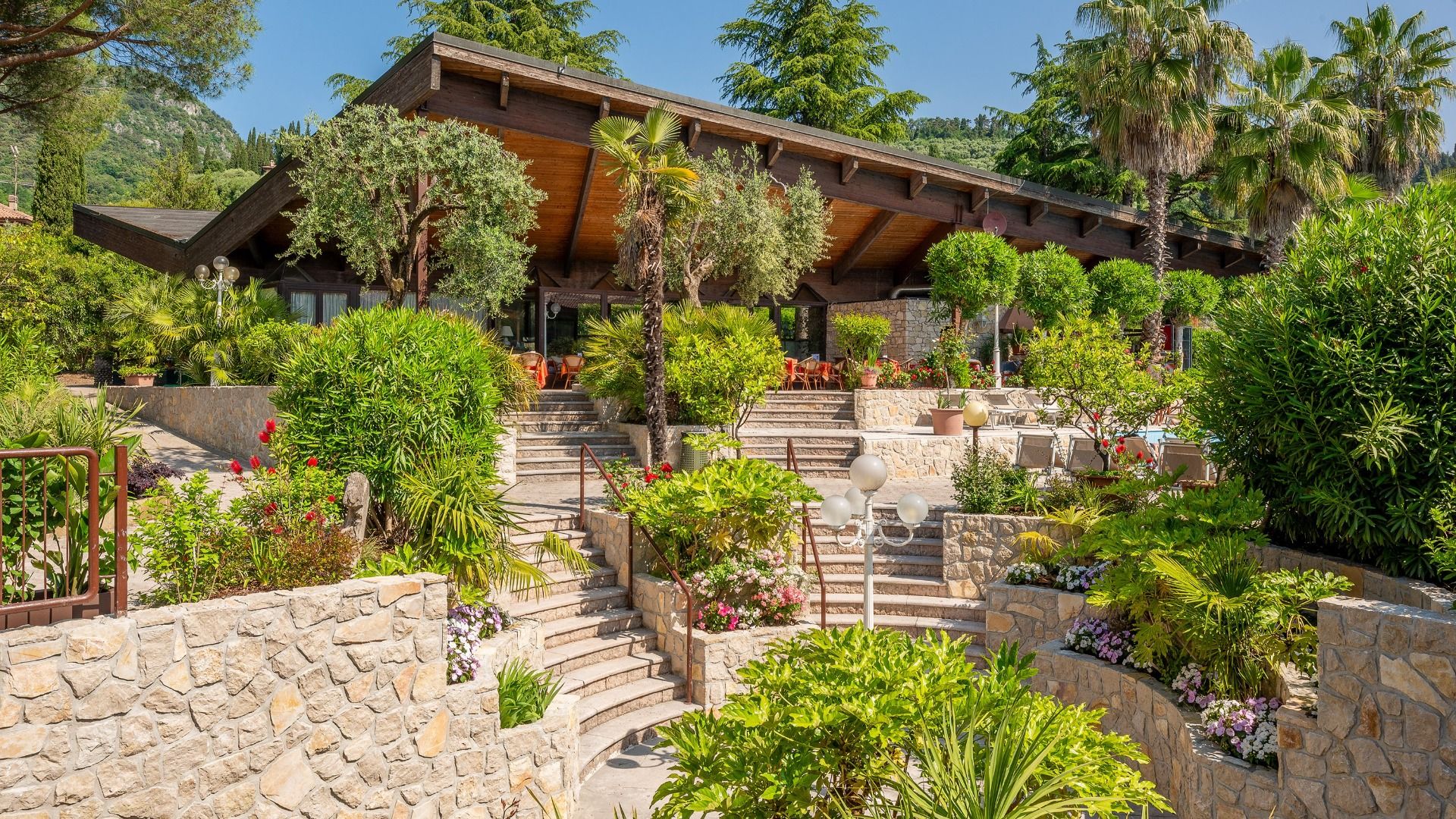 Ein modernes Haus mit einem großen Holzdach liegt hinter terrassenförmig angelegten Steinstufen inmitten von üppig grünen Bäumen, Palmen und bunten Blüten - und erinnert an das Sporthotel Olimpo am Gardasee unter einem klaren blauen Himmel.