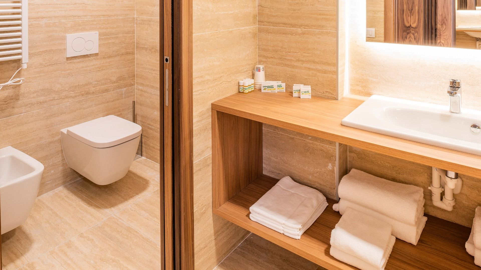 Das moderne Badezimmer im Sporthotel Olimpo verfügt über beigefarbene Fliesen, eine Wandtoilette, ein Bidet, einen hölzernen Waschtisch mit weißem Waschbecken, offene Regale mit gefalteten Handtüchern, Toilettenartikel auf dem Tresen und eine helle, saube