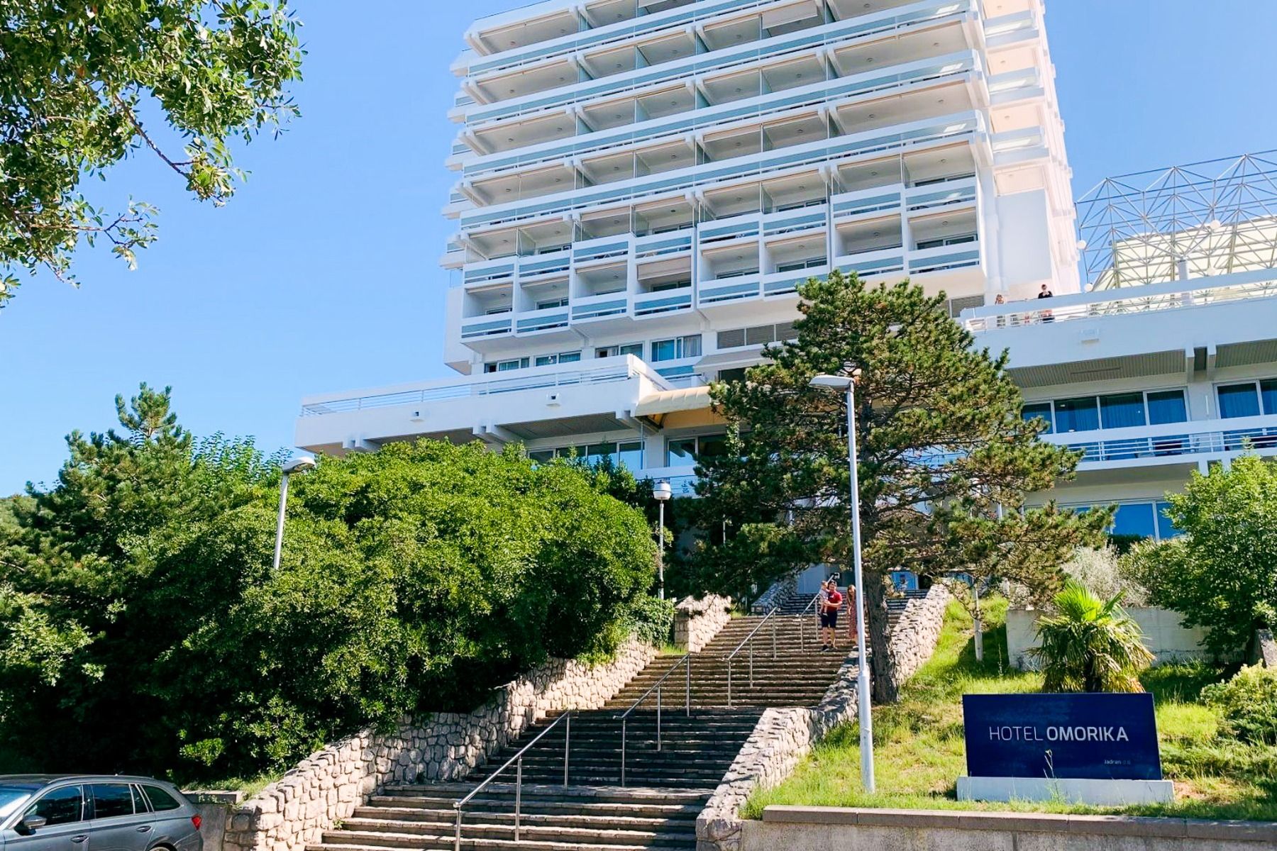 Hinter üppig grünen Bäumen und Sträuchern erhebt sich das hohe, moderne, weiße Gebäude des Hotels Omorika. Eine lange Steintreppe führt hinauf zum Eingang, an dessen Vorderseite ein blaues „Hotel Omorika“-Schild angebracht ist.