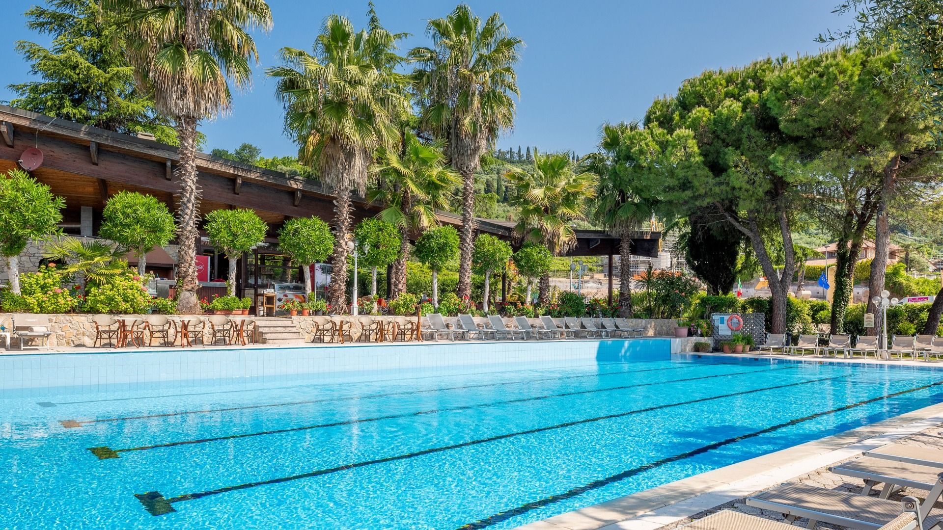 Das Sporthotel Olimpo verfügt über einen großen Außenpool mit Bahnen, Liegestühlen und Palmen inmitten von Grün in der Nähe von Tennisplätzen und dem Gardasee, direkt neben einem Holzgebäude.