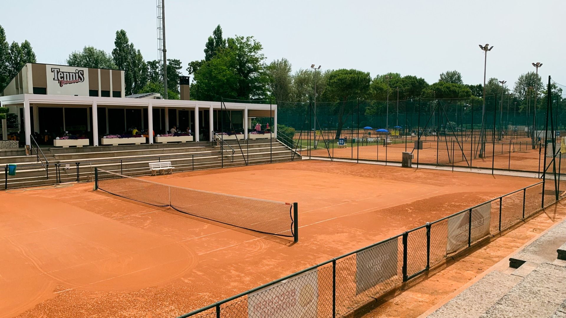 Sandplatz-Tennisanlage mit Clubhaus und Zuschauerrängen, umgeben von Bäumen und weiteren Tennisplätzen im Hintergrund.
