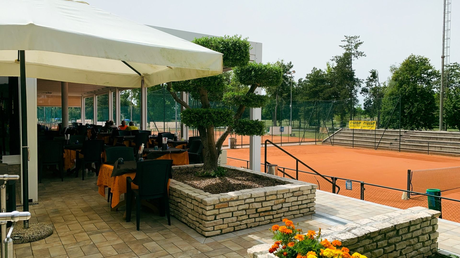 Terrasse des Clubhauses mit Sitzplätzen und Blick auf den angrenzenden Sandplatz-Tennisplatz, dekoriert mit Blumen und Sonnenschirmen. 