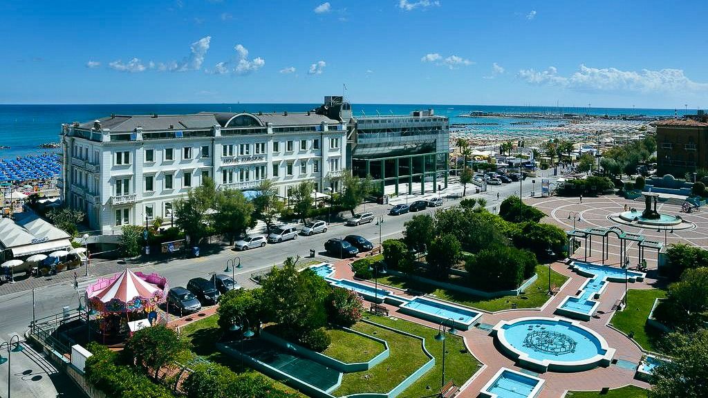 Luftaufnahme der Hotelanlage Kursaal an der Strandpromenade mit Parkanlage, Brunnen und Blick auf das Meer.
