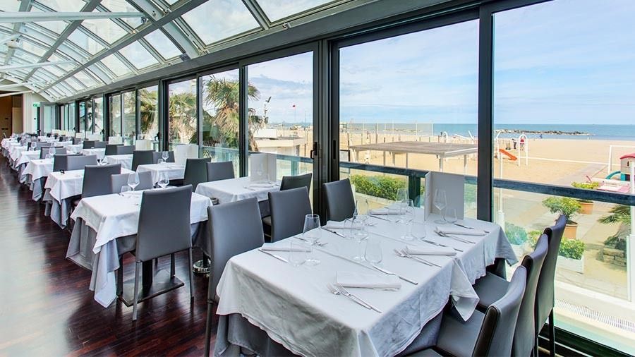 Panorama-Restaurant mit Meerblick, gedeckte Tische und große Fensterfront mit Blick auf den Sandstrand.