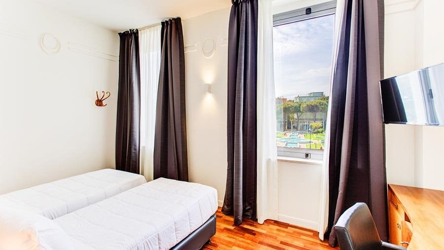 Modernes Doppelzimmer mit zwei Einzelbetten, helle Vorhänge, Holzfußboden und Fenster mit Blick auf die Parkanlage.