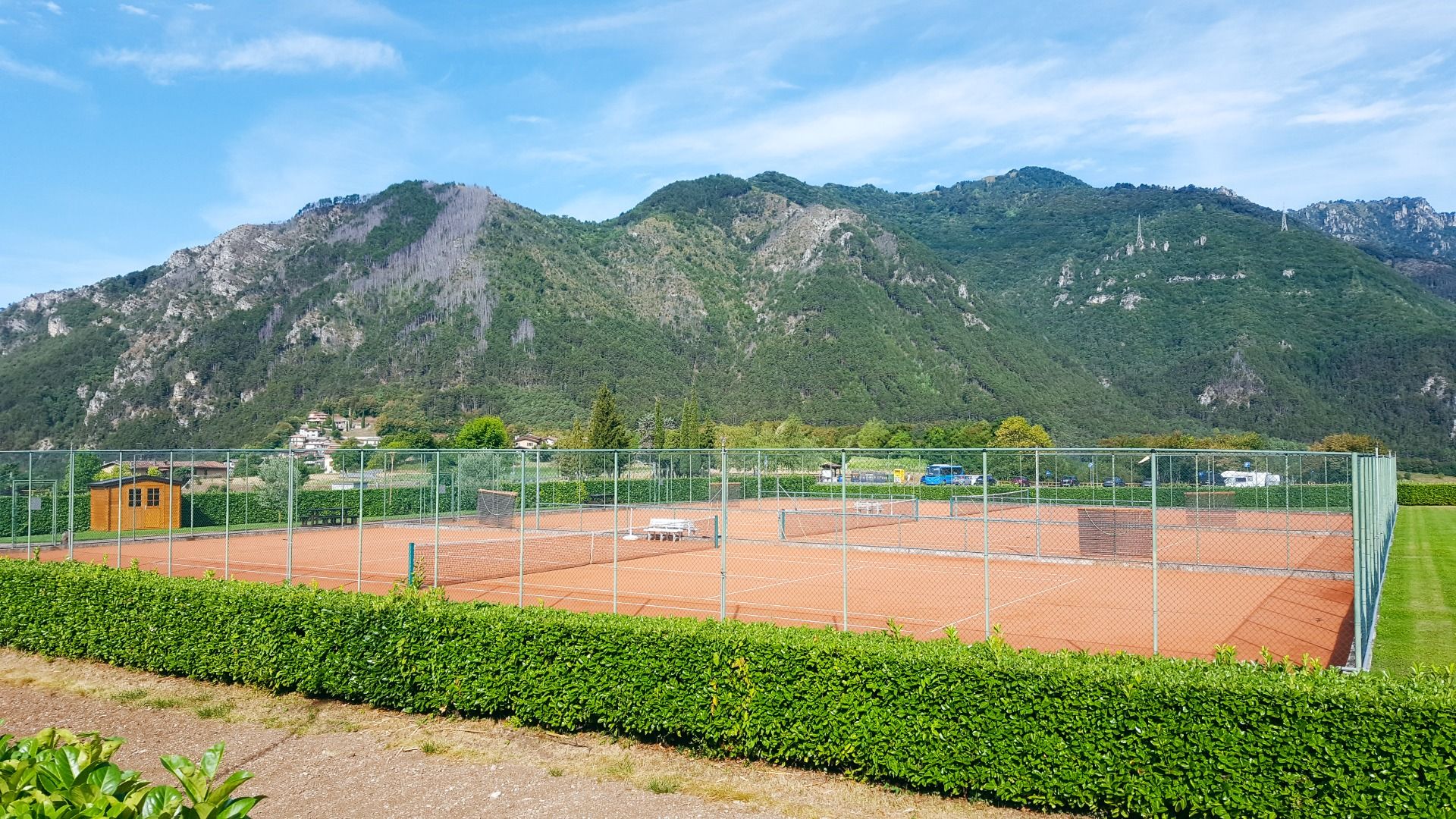 Tennisplätze mit rotem Sand, umgeben von Zaun und grüner Hecke, mit Bergen im Hintergrund.