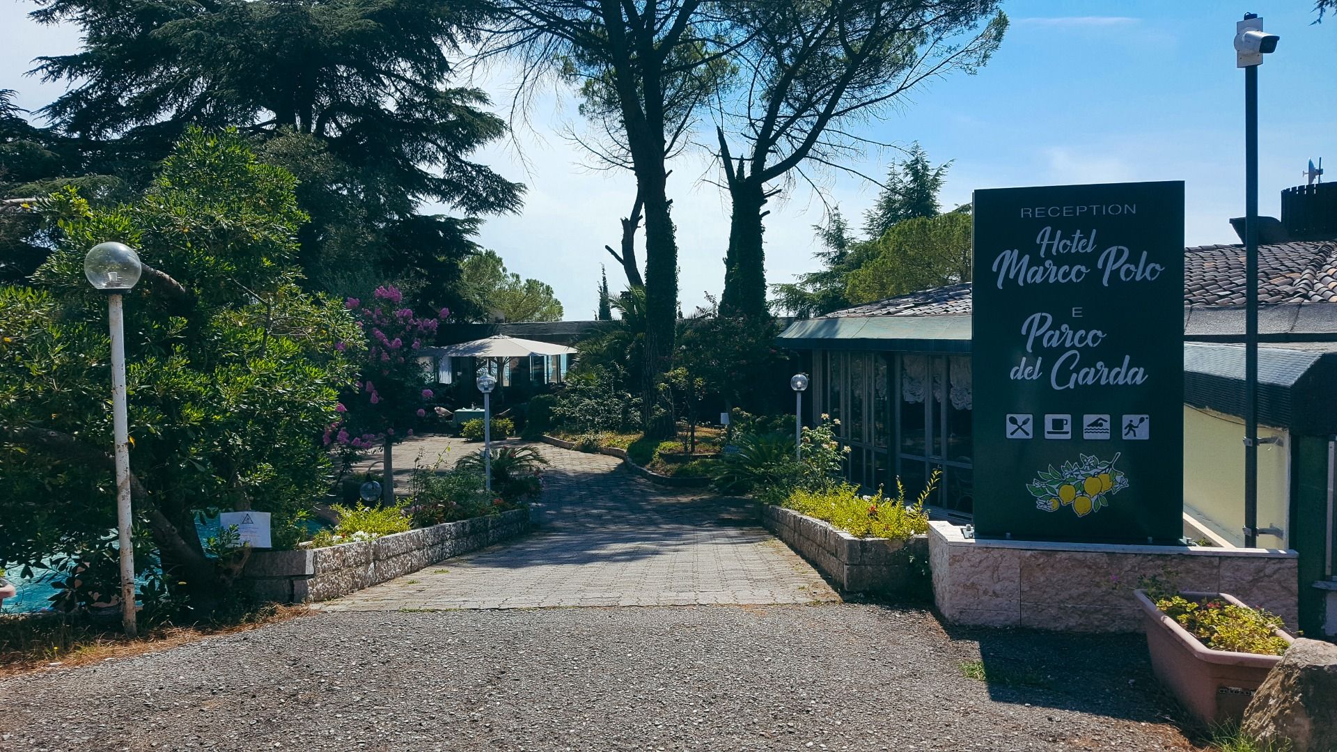 Eingangsbereich des Hotels Marco Polo, von Bäumen und Pflanzen umgeben, mit einem Weg zur Rezeption und einem großen Schild mit Hotelnamen.