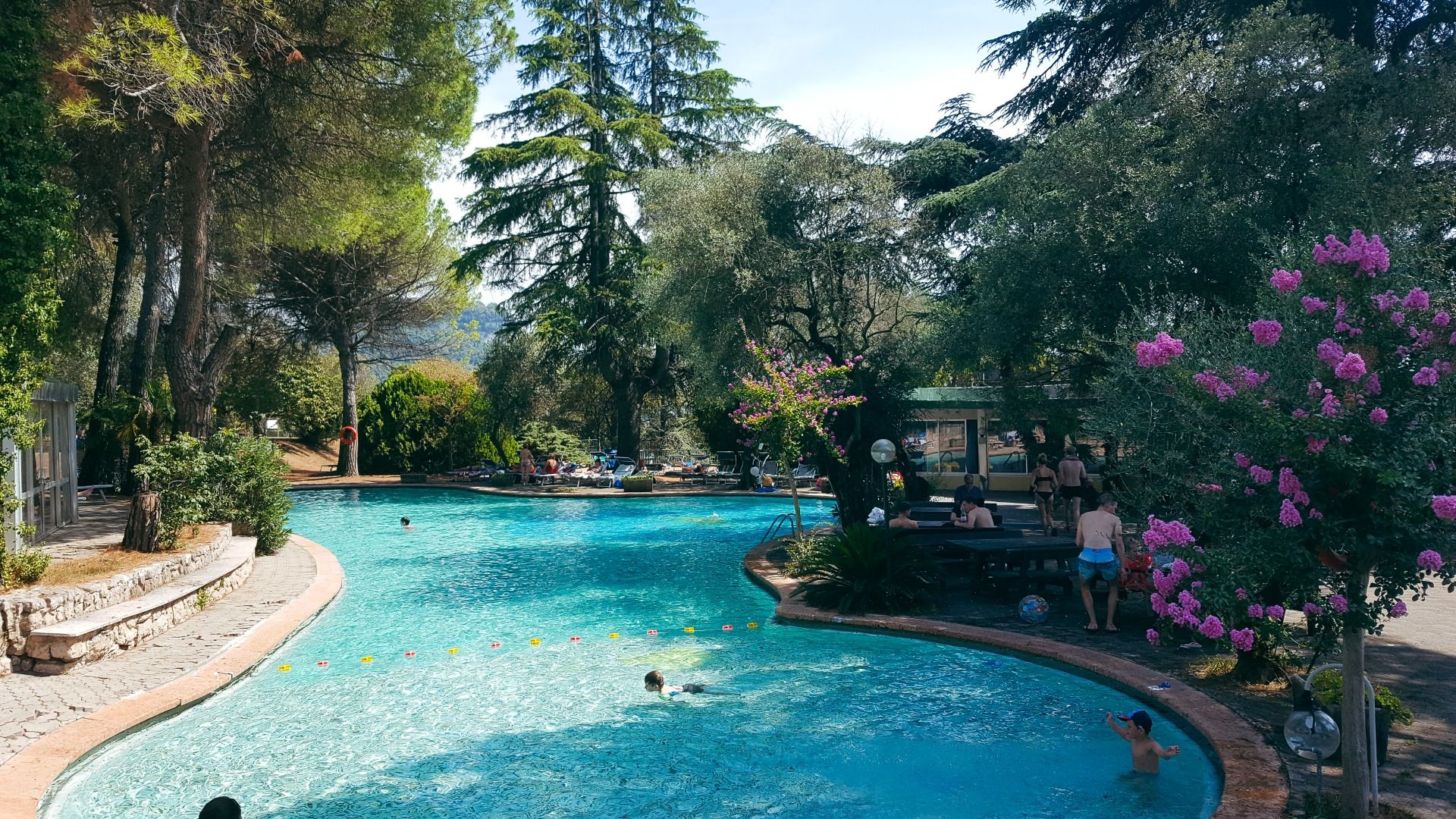 Swimmingpool inmitten einer Gartenlandschaft mit großen Bäumen und blühenden Pflanzen, Gäste schwimmen und entspannen am Beckenrand.