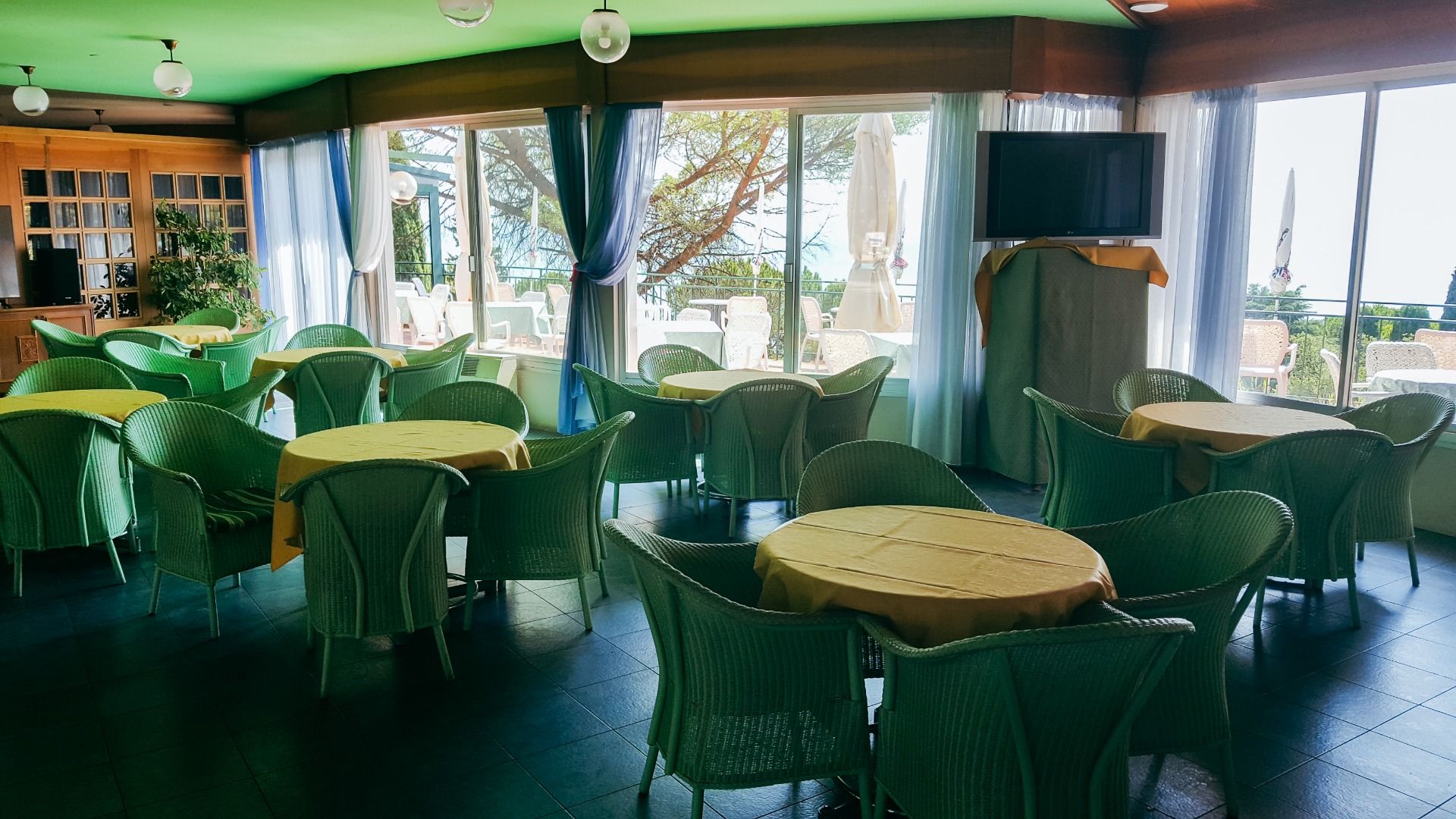 Innenbereich einer Lounge mit grünen Korbmöbeln und gelben Tischdecken, große Fenster mit Blick auf die Terrasse.
