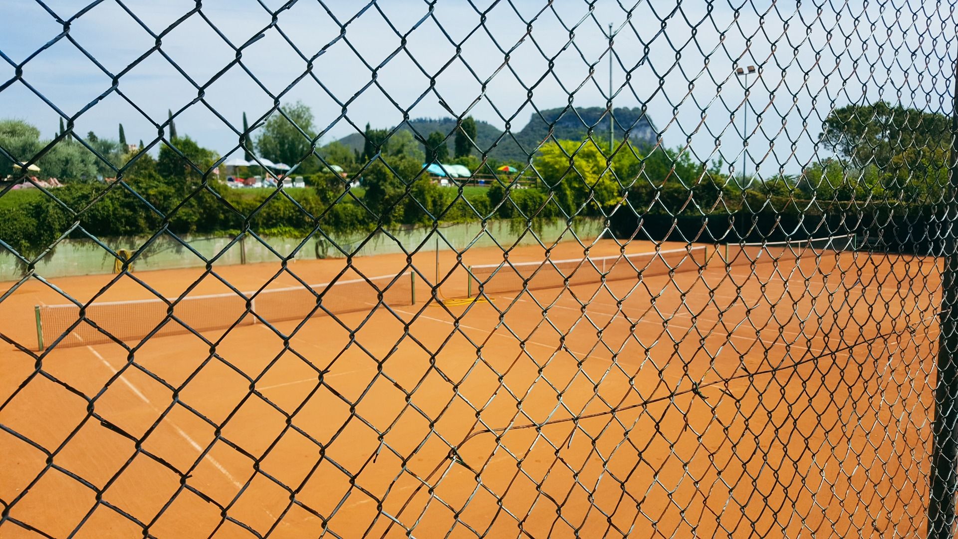 Tennisplatz mit rotem Sandbelag, fotografiert durch einen Maschendrahtzaun, im Hintergrund Hügel und grüne Landschaft.