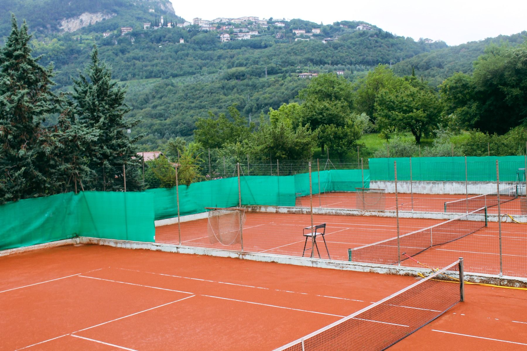 Tennisplätze mit rotem Sandbelag, grünem Sichtschutz und bewaldeter Berglandschaft im Hintergrund.