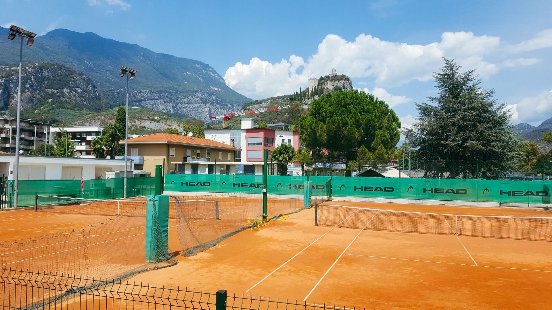 Sand-Tennisplatz mit Flutlicht, Bergen und einer Burgruine im Hintergrund.