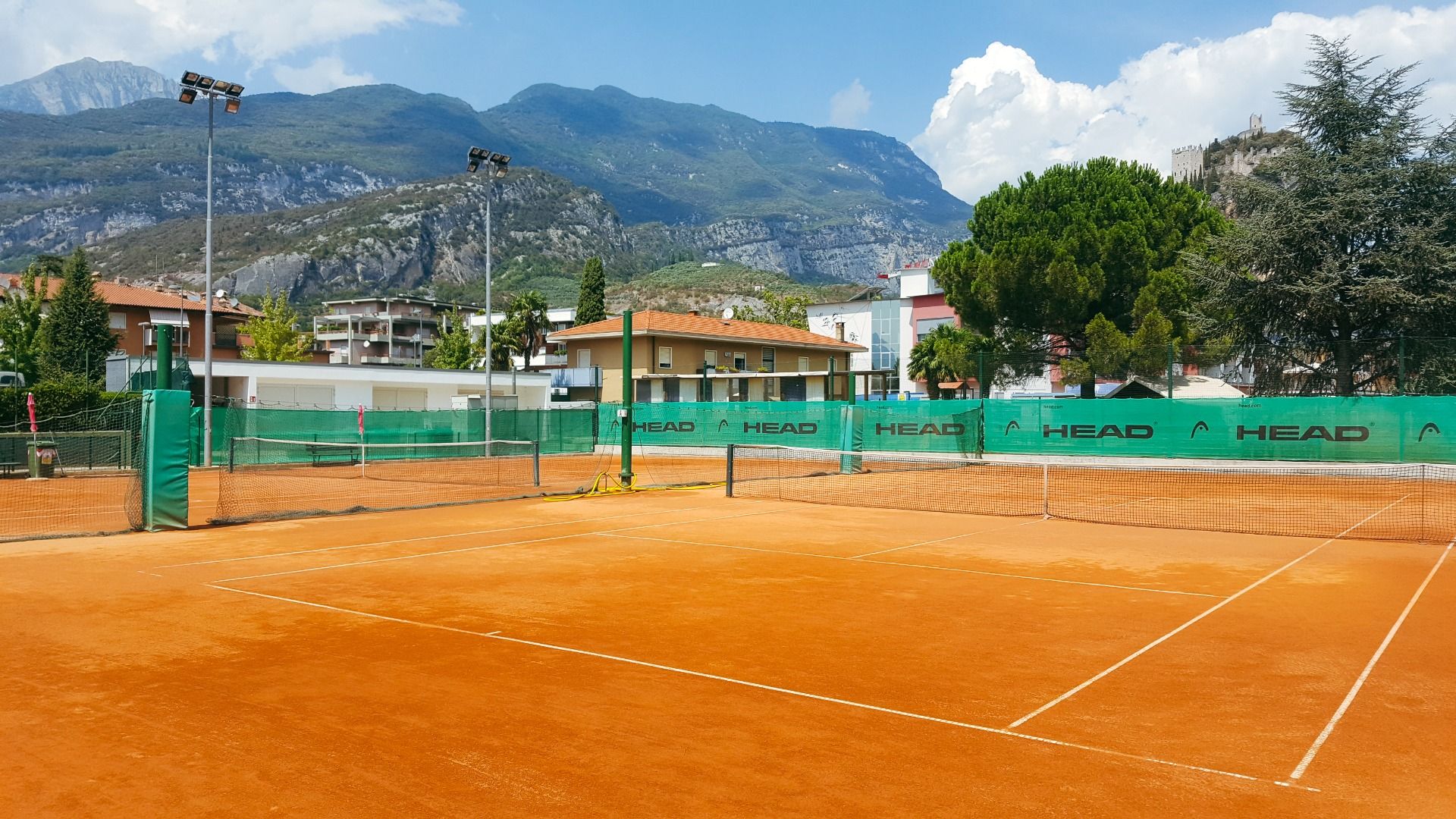 Sand-Tennisplatz mit umliegenden Gebäuden, Bergen und Flutlicht.